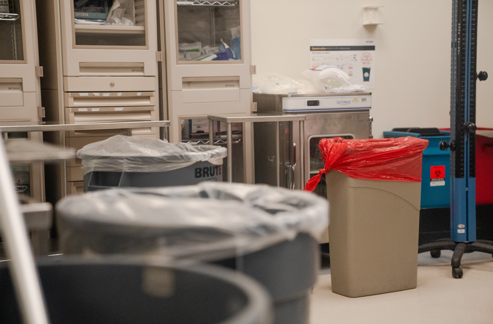 In an empty operating room, a large trash can with a clear plastic bag is seen in the foreground and a smaller can with a red bag in the background.